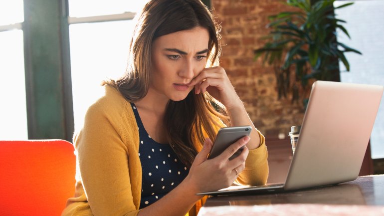 Eine Frau sitzt am Laptop und schaut angespannt auf ihr Smartphone.