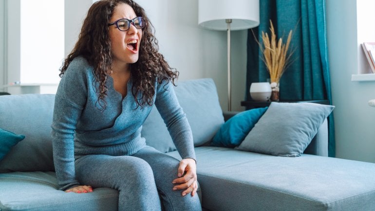 Frau mit Synovitis sitzt auf Sofa und fasst sich an schmerzendes Knie.