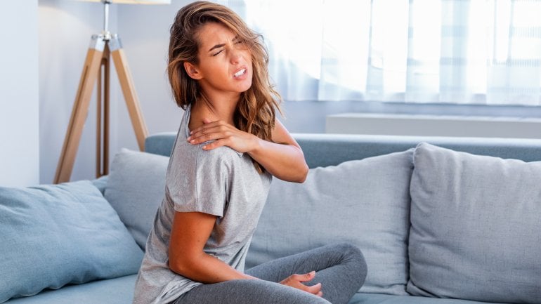 Frau mit Kalkschulter sitzt auf dem Sofa und fasst sich an schmerzende Schulter.