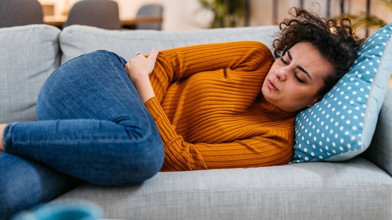 Frau liegt mit starken Schmerzen auf dem Sofa und hält sich den Bauch