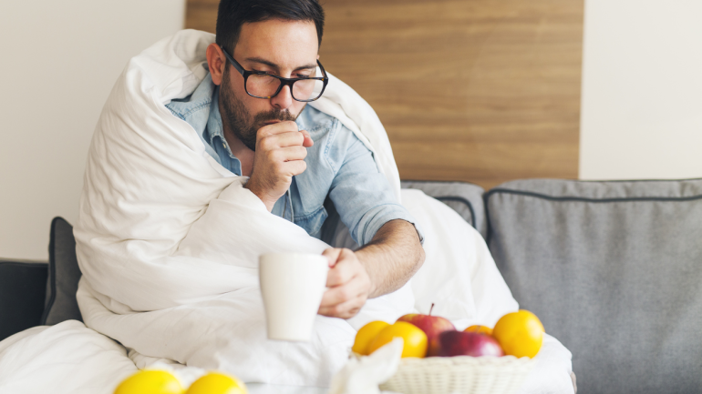 Akute Bronchitis: Ein Mann sitzt in einer Decke gehüllt auf dem Sofa und hustet.