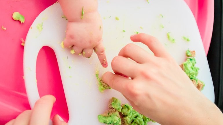 Man sieht eine Erwachsenen- und eine Babyhand mit Brothäppchen mit Avocado