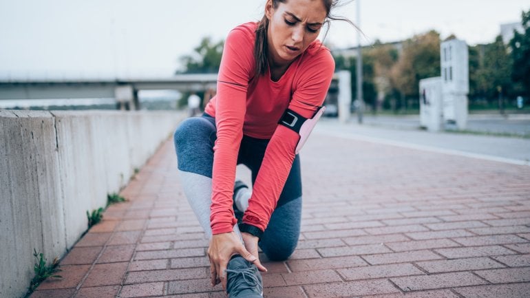 Frau macht Sport, zieht sich einen Bänderriss zu und fasst sich an schmerzenden Knöchel.