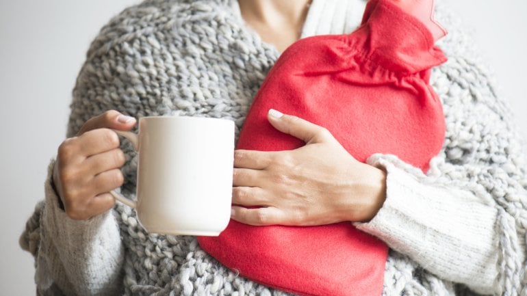 Eine Frau hält eine Tasse in der rechten Hand und eine rote Wärmflaschen im linken Arm.