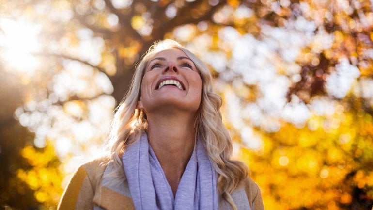 Eine Frau steht im Herbst in einem Wald und schaut lächelnd in die Bäume, die von der Sonne angestrahlt werden. 