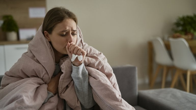 Man sieht eine hustende Frau, die in einer dicken Decke eingewickelt auf der Couch sitzt