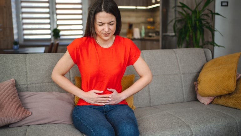 Frau mit Colitis ulcerosa sitzt auf Sofa und fasst sich an schmerzenden Bauch.