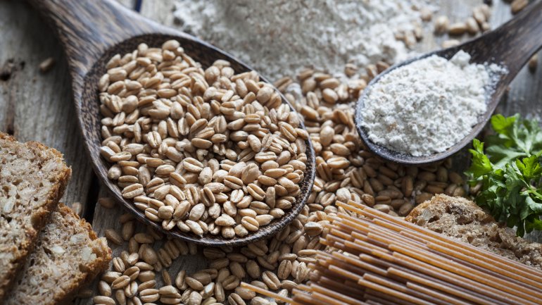 Zu sehen ist ein Holzlöffel mit Dinkelkörnern, daneben Mehl, Brot und Nudeln aus Dinkel