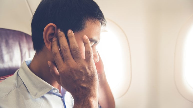 Mann im Flugzeug hat Druck auf den Ohren und fasst sich mit Händen an den Kopf.