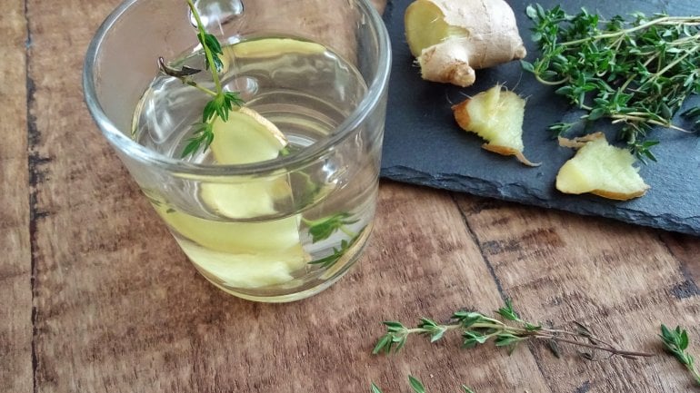 Auf einem Holztisch liegt ein Schneidebrett mit Thymianzweigen und angeschnittener Ingwerwurzel, daneben steht ein Glas mit Thymian-Ingwer-Tee.