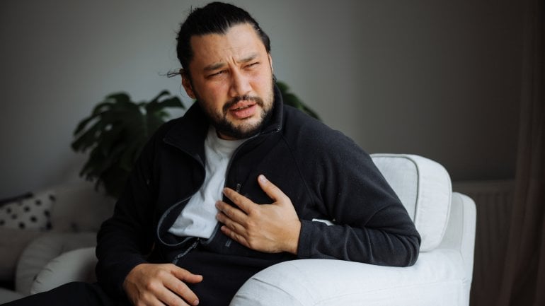 Junger Mann mit Herzmuskelentzündung sitzt auf Couch und hält sich Hand auf schmerzende Brust.