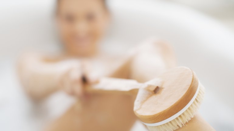 Eine Frau liegt in der Badewanne und streicht mit einer Badebürste über ihren Unterschenkel.