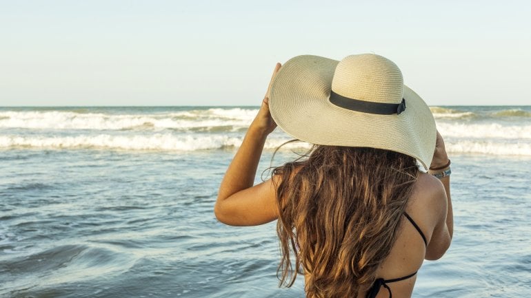 Eine Frau steht in Bikini und mit Sonnenhut im Meer.