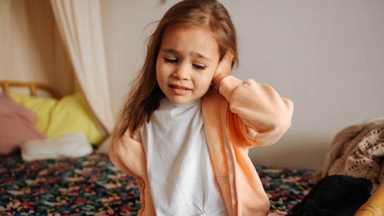 Kleines Mädchen mit Mastoiditis sitzt auf Bett und fasst sich ans Ohr.