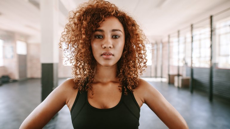 Eine Frau mit dunklen Locken und sportlichem Outfit blickt gerade in die Kamera.