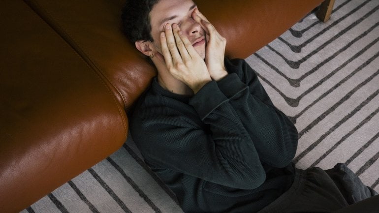 Junger Mann mit Migräne sitzt vor der Couch und hält sich Hände an den schmerzenden Kopf.
