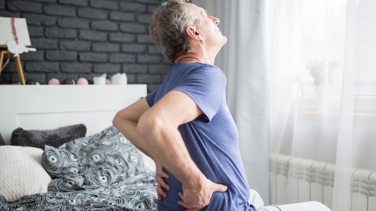 Ein Mann sitzt nach dem Aufwachen auf dem Bett und hält sich den schmerzenden unteren Rücken