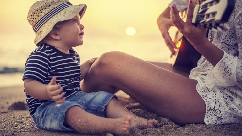 Ein Baby lauscht am Strand seiner Mutter, die Gitarre spielt.