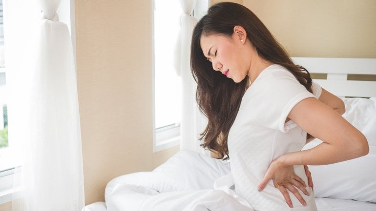 Eine jugne Frau sitzt auf dem Bett und fasst sich an die Nieren. Sie hat Schmerzen