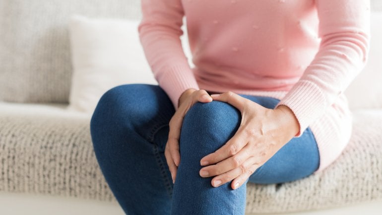 Eine Frau sitzt auf dem Sofa und umfasst ihr schmerzendes Knie.