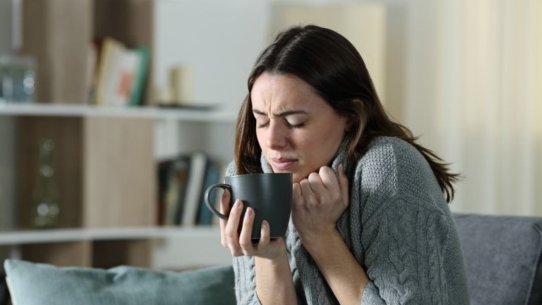 Frau sitzt auf Sofa mit Decke und Tee und hat Schüttelfrost.