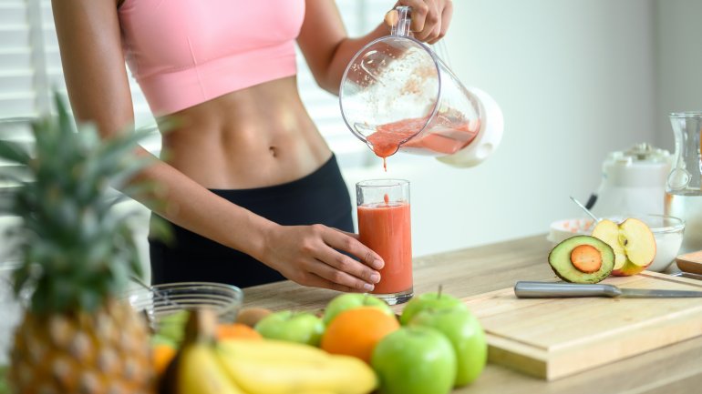 Eine Frau mit Sixpack steht in der Küche und bereitet sich einen Smoothie zu. 