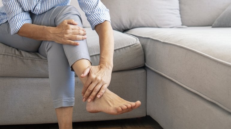 Frau sitzt auf Sofa und berührt ihr Bein aufgrund von Taubheitsgefühlen.