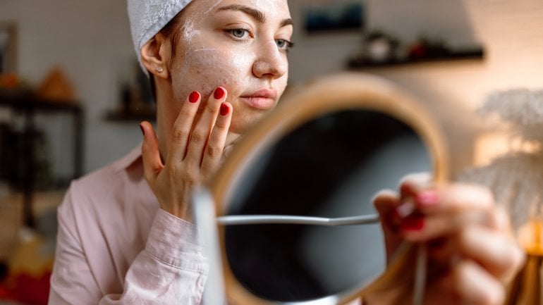 Eine junge Frau mit Pickeln schaut in den Spiegel und trägt eine Maske auf