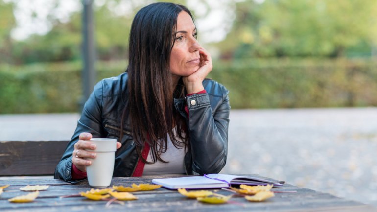 Eine Frau mittleren Alters sitzt an einem Herbsttag draußen an einem Tisch, vor sich ein aufgeschlagenes Notizbuch, in der rechten Hand einen Kaffeebecher haltend und mit der linken Hand ihren Kopf stützend.