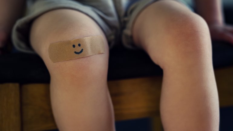 Zu sehen ist das Knie eines Kindes mit Pflaster, auf dem ein lächelnder Smiley aufgemalt ist