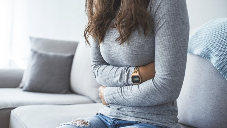 Eine Frau sitzt auf dem Sofa und hat die Arme über den Bauch gelegt.